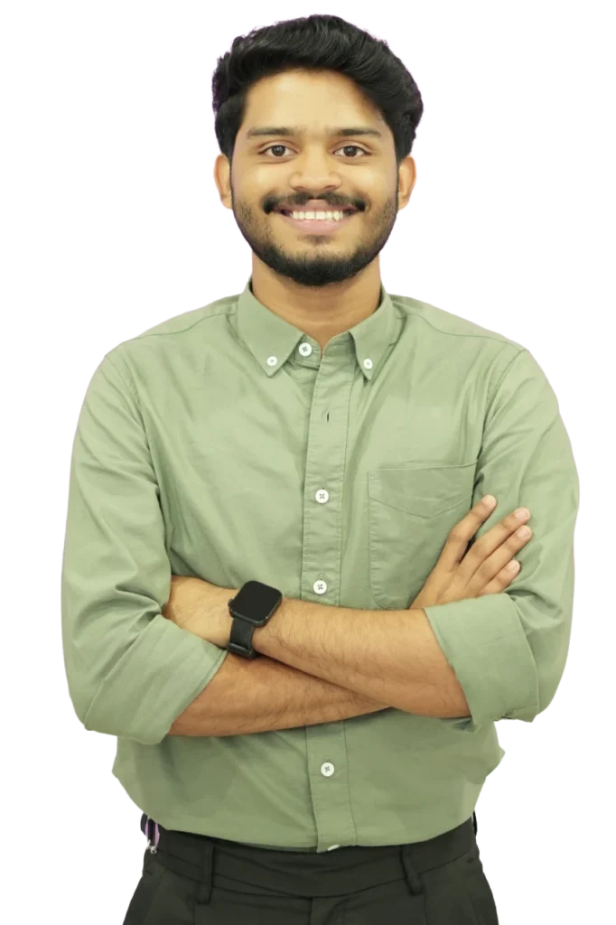 Smiling young man with short black hair and a trimmed beard, wearing a light green button-down shirt and a smartwatch, standing with arms crossed against a plain background.